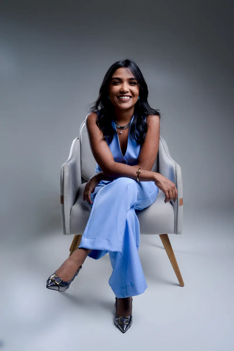 Samara Farias sorrindo de frente, de macacão azul-claro, sentada numa poltrona cinza