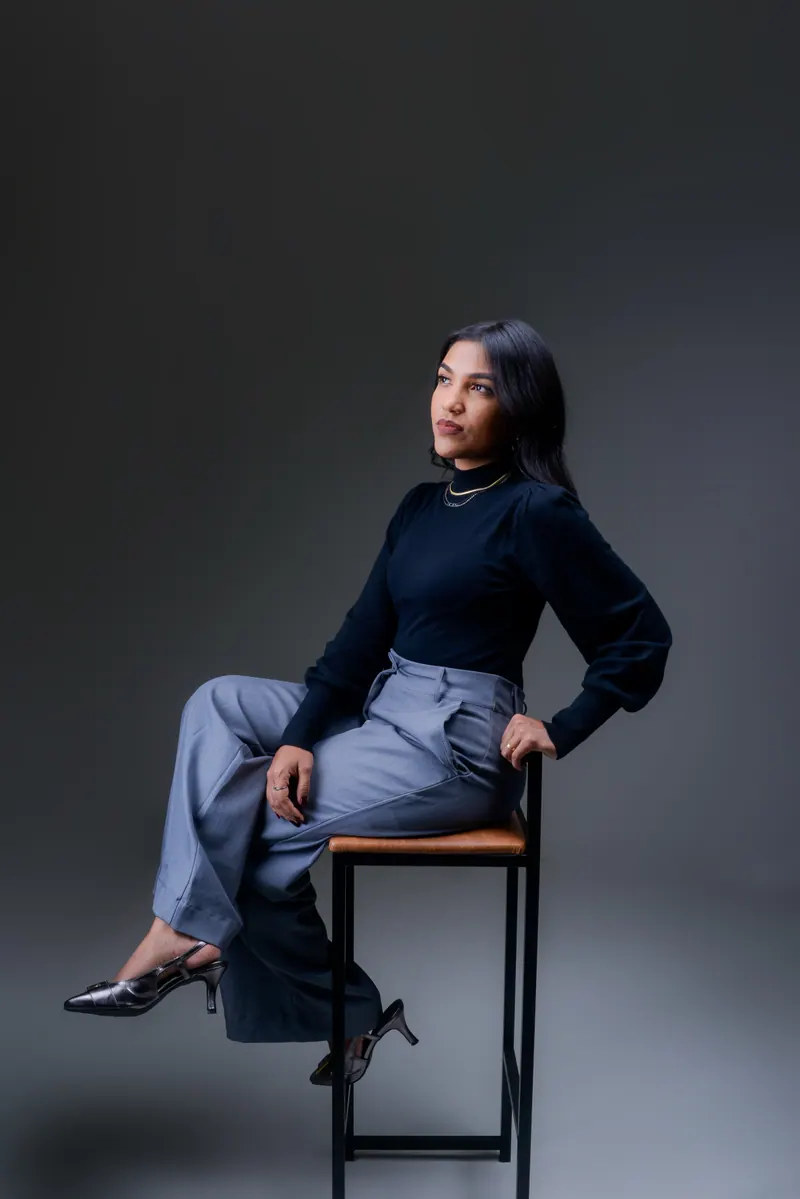 Samara Farias de blusa azul-marinho e calça cinza-azulada, sentada numa banqueta de assento caramelo, em fundo cinza